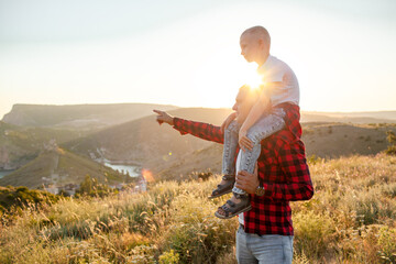 Fototapeta na wymiar Happy family spending time together in the nature, father holding son on shoulder, have fun, playing with kid in vacation against the background the grass, mountain and sunset