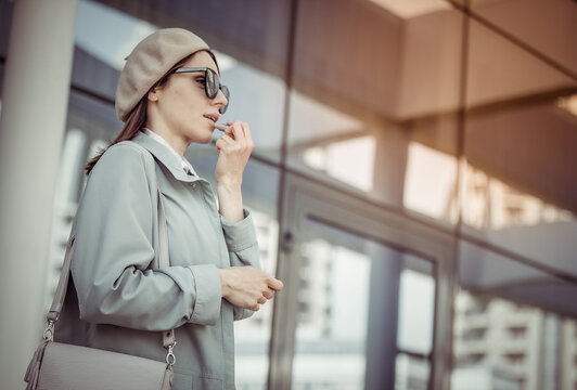 Young stylish woman in sunglasses and beret paints her lips against the background of the windows of the business center. Fashion, beauty, lifestyle