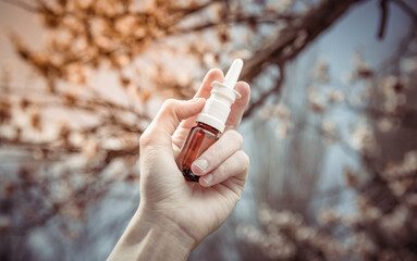 Hand holds nasal spray on background of blossoming tree. Spring exacerbation of allergies