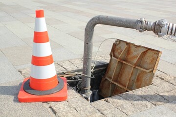 Open manhole, construction traffic warning cone and metal pipe on sidewalk. Concept of repair of...