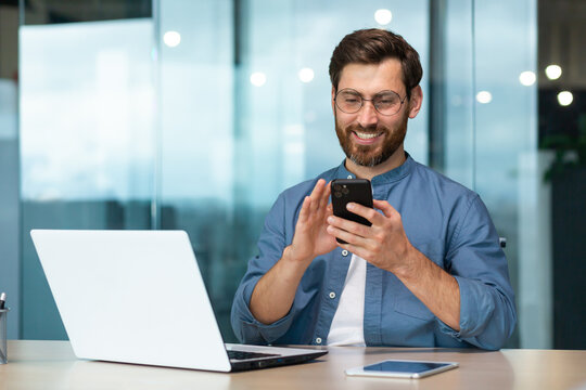 Successful and happy businessman with beard works in modern bright office, male boss uses laptop, business owner in casual shirt holds phone, browses internet pages