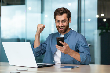 Businessman celebrating success and triumph, man in shirt working inside modern office with laptop holding phone reading message, business owner received good news notification online - obrazy, fototapety, plakaty