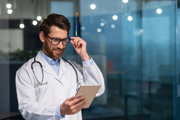 Cheerful and smiling senior doctor in glasses and medical gown reading message from tablet computer, man working inside modern clinic