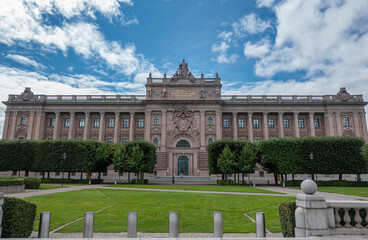 Sweden, Stockholm - July 16, 2022: East Facade, Beige stone F&ouml;rsvarsutskottet or Riksdagshuset or Parliament behind green park on its east side under blue cloudscape. Statues on gable