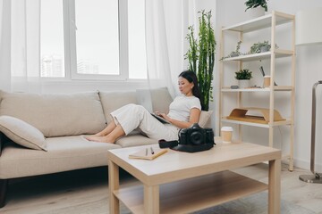 Asian woman photographer lying at home on the couch and reviewing photos from the camera in laptop for color correction retouching, work as a freelance business photographer for yourself home office