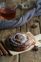 A sweet bun with chocolate and cinnamon lies on a board on a wooden table. Behind is a towel and a cup of tea. Nearby are cinnamon sticks and star anise