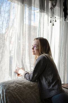 beautiful young woman looks thoughtfully and dreamily out the window at home. Loneliness and melancholy in women