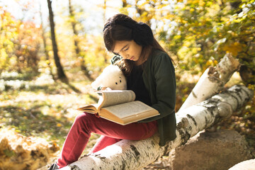 A girl reads a book to her soft toy. A girl and her toy are reading a book. Child reading a book. Children's books. A child reads books in the autumn forest.