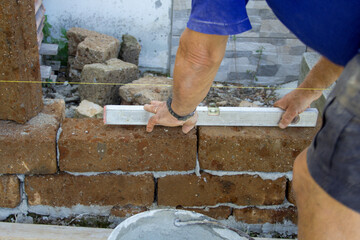Image of the hands of a bricklayer who checks the regularity of the plane of the tuff brick wall he is building with a level.

