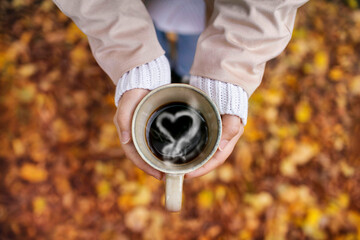 Person holding in hands a big mug of hot black coffee in autumn scenery; In love with autumn