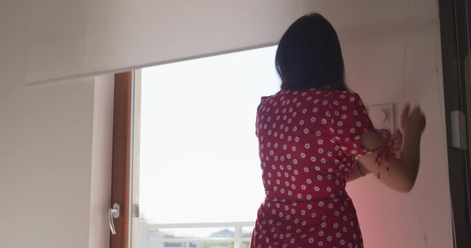 A happy young woman in red dress raises a white roller blinds in the morning. A beautiful girl at home.
