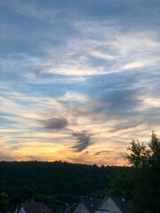 Fototapeta na wymiar illustration d'un ciel avec des nuages gris et le coucher du soleil et en premier plan des arbres et toitures de maison
