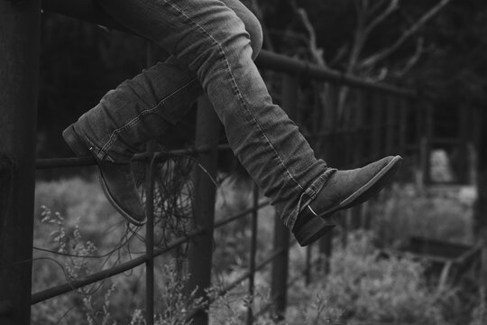Western person sitting on fence with cowboy boots on farm in black and white.