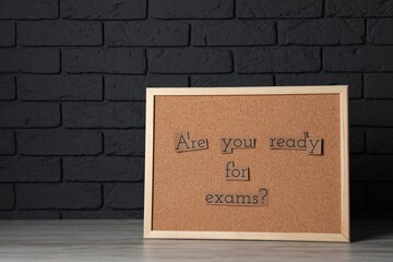 Obraz premium Cork board with phrase Are You Ready For Exams? on wooden table near black brick wall. Space for text