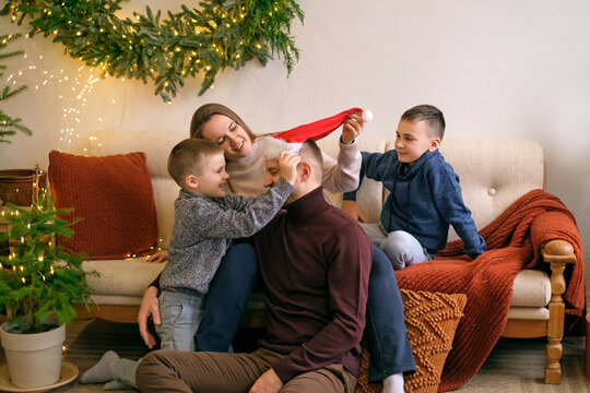 Sons And Mom Dress Up Dad In Santa Claus. Children Have Fun With Their Parents On Christmas Eve.
