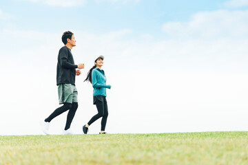 青空の見える公園でランニングするスポーツウェアを着たアジア人の男女（走る）
