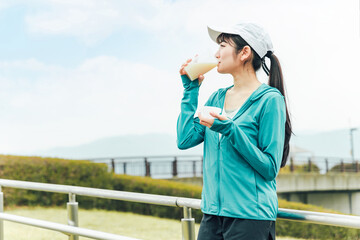 ボディメイクのため公園でプロテインを飲みながらトレーニングするスポーツウェアのアジア人女性
