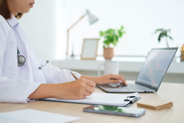 Cropped shot of doctor in white uniform using laptop and filling up medical recommendations in a clinic office.