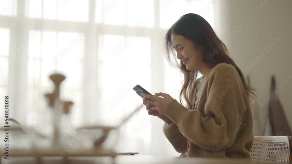 Poster Happy young asian woman relaxing at home. Asia female standing at counter kitchen and using mobile smartphone