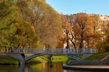 Pedestrian bridge across the canal in the city autumn park, no people. Trees in colorful autumn foliage next to a pond.