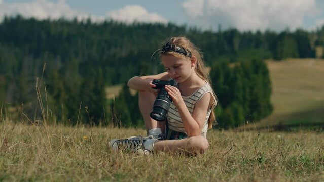Young photographer watching results of work, sitting on mild grass hill. Girl relaxing and feeling warm wind currents, spending leisure time outdoor