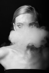 Fashion, style and make-up concept. Close-up black and white studio portrait of beautiful blonde woman blowing smoke from mouth. Model looking to camera with seductive look. Hair in camera focus