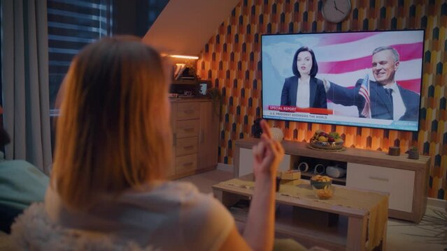 Couple sitting on sofa in living room, watching politics TV news and speech of President of United States, showing middle finger, eating pizza. Spending evening at home together. TV live broadcasting.