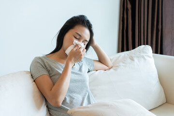 Sick young Asian woman sitting on couch blowing her nose on a tissue conceptual of healthcare, seasonal flu, allergic rhinitis or allergy reaction in hay fever. Closeup, copy space