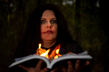 Black haired witch holding burning book in her hands in forest.
