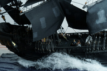 Pirate ship model Black pearl made of plywood, with miniature figures of pirate sailors, close-up details. Ship sails on wave. Sailors on deck pulling ropes, partially blurred. Patched sails belly out