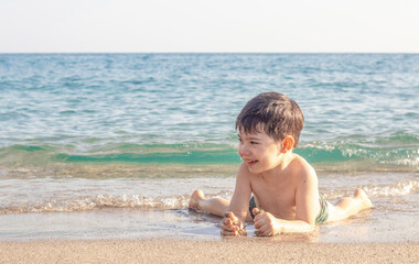 little boy kid having fun in sea ocean  water lying on sands beach splashing hitting waves with hand putting sand on legs belly tummy playing smiling.happy child enjoy vacation sunny summer day