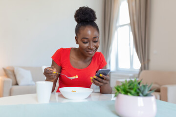 Beautiful young arfican american woman texting on smart phone at home. Cheerful lady eating corn flakes, drinking morning coffee and looking at her mobile phone