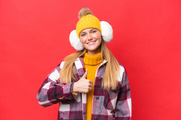 Young beautiful woman wearing winter muffs isolated on red background giving a thumbs up gesture