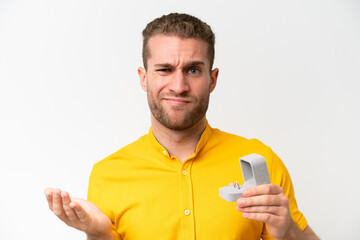 Young man holding a engagement ring isolated on white background making doubts gesture while lifting the shoulders