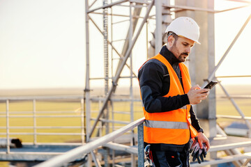 A serious industry worker is receiving important message on the phone while standing on high metal construction.