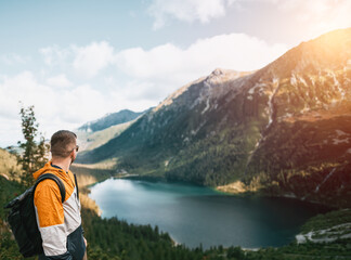 Fototapeta na wymiar Man traveler smiles with beatufil mountain lake and forest in the background. Concept of outdoor adventures and hiking. Bearded young caucasian man wears backpack and sport outfit in the mountains.