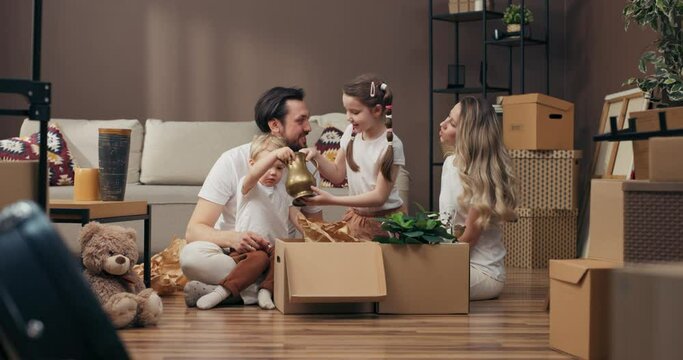Moving in process concept happy young family sitting on floor in own new apartment with carton boxes full of stuff curious son taking vase putting on new table decorating furnishing new home .