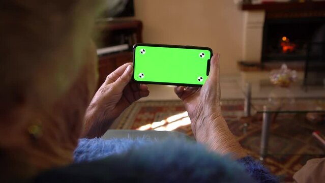 Wrinkled hands of the old woman holds a smartphone with green screen for your mockup. Close up elderly hands of caucasian grandma showing blank phone for new mobile app or website.