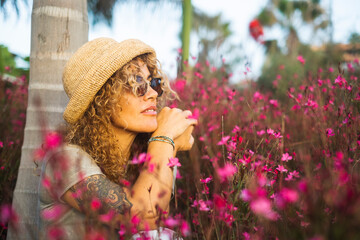 Side portrait of cute young woman smiling and enjoying garden flowers and nature feeling in spring season. Happy female people sitting in the middle of pink flowers park. Concept of people and nature