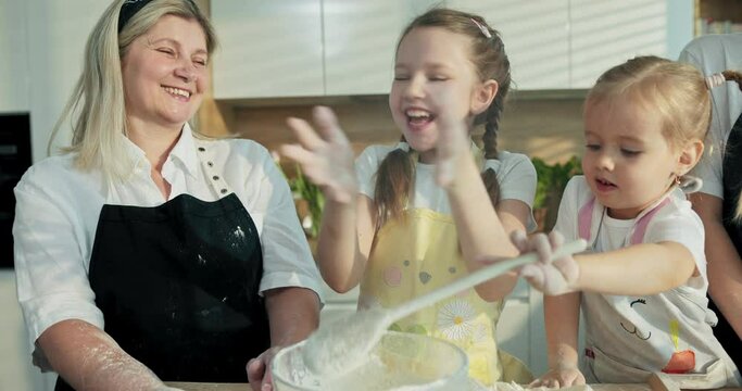 Older granddaughter clepping in hands with flour luaghing having fun with grandmoother preschooler sister and young mother in modern kitchen while little girl trying to help with big spoon in hands.