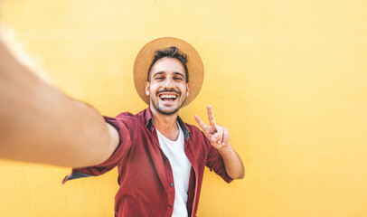 Handsome man wearing straw hat smiling at camera on a yellow background - Isolated happy guy taking selfie picture with smart mobile phone device