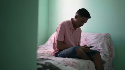 One young hispanic man looking at cellphone device sitting on bed inside bedroom