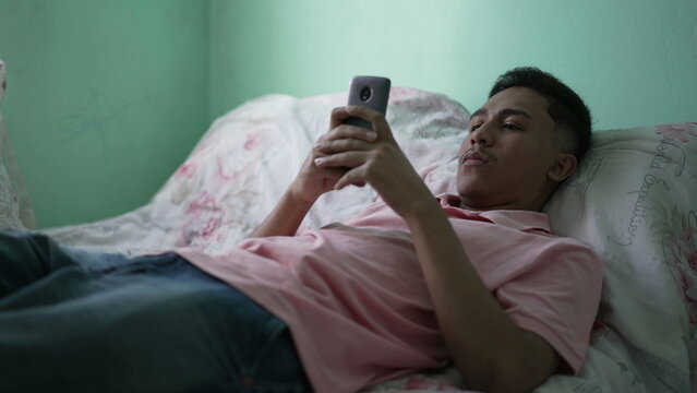 One Casual Hispanic Man Holding Phone In Bed. Relaxed Person Lying Down Looking At Smartphone Device Screen