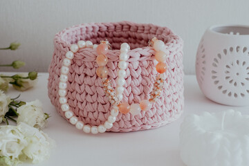 Obraz na płótnie Canvas Pearl necklace and beads in pink crocheted basket, white candle, candleholder and lisianthus bouquet on white table in white interior. Cozy home atmosphere, Handicraft advertisement