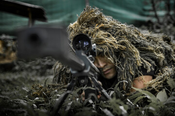 Man in a ghillie suit hidden in the forrest, aiming through a scope on a rifle. May both represent...