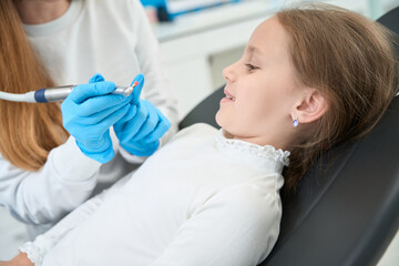 Pediatric dentist telling about teeth-polishing procedure to smiling little girl