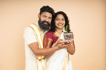 Happy South Indian couple holding jar filled with money saving coins and plant growing isolated on...