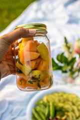 Black woman holding a glass jar of fruit salad while enjoying a luxury sunset picnic in the park 
