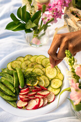 Black woman holding a cucumber slice from crudites platter while enjoying a luxury sunset picnic in the park  - obrazy, fototapety, plakaty
