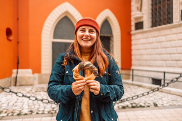 Fall tourist woman in a bright hat and autumn jacket holding baked obwarzanek traditional polish cuisine snack bagel on old city Market square in Krakow 